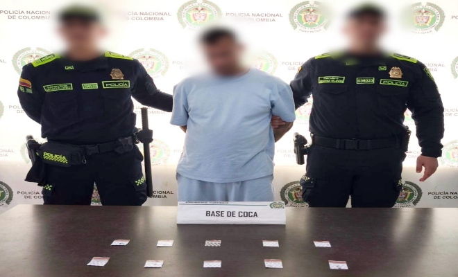 Dos funcionarios policiales con un ciudadano capturado frente a pendón institucional y delante una mesa, sobre la cual esta en material incautado