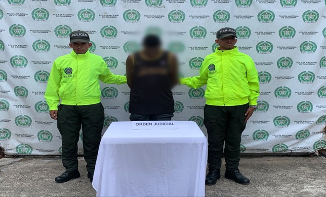 Dos uniformados de la Policía Nacional presentan a una persona capturada mediante orden judicial durante un procedimiento realizado en Quibdó. La persona se encuentra ubicada entre los funcionarios policiales frente a una mesa cubierta con un mantel blanco, sobre la cual se observa un aviso con la leyenda “orden judicial”. Al fondo se aprecia un pendón institucional con los emblemas de la Policía Nacional de Colombia.