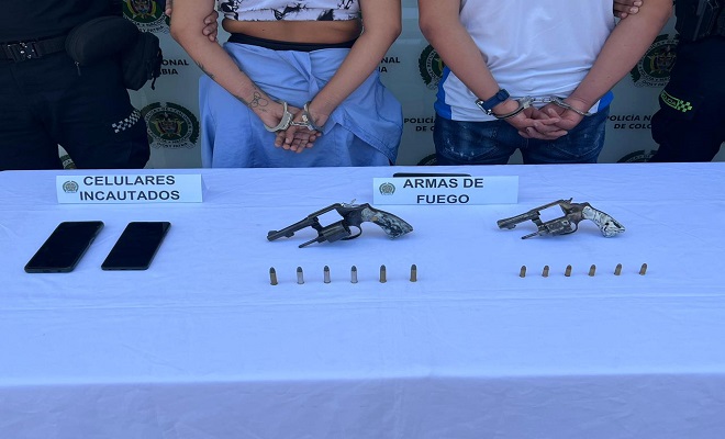 Elementos incautados, entre ellos dos armas de fuego y dos celulares, de fondo dos personas capturadas y dos funcionarios de la Polic&iacute;a Nacional