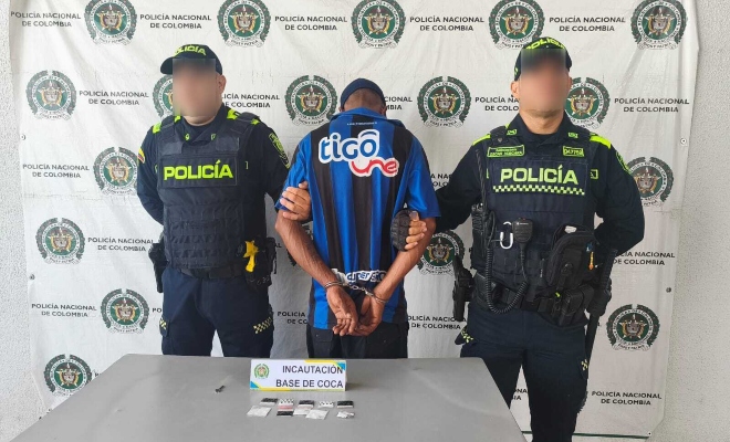 Dos funcionarios policiales junto a un ciudadano capturado frente a pend&oacute;n institucional frente a ellos una mesa sobre la cual est&aacute;n las sustancias estupefacientes incautadas 