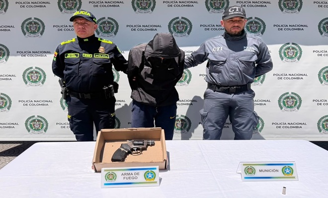 un policía, una persona del CTI, un capturado, una mesa, un arma de fuego incautada 