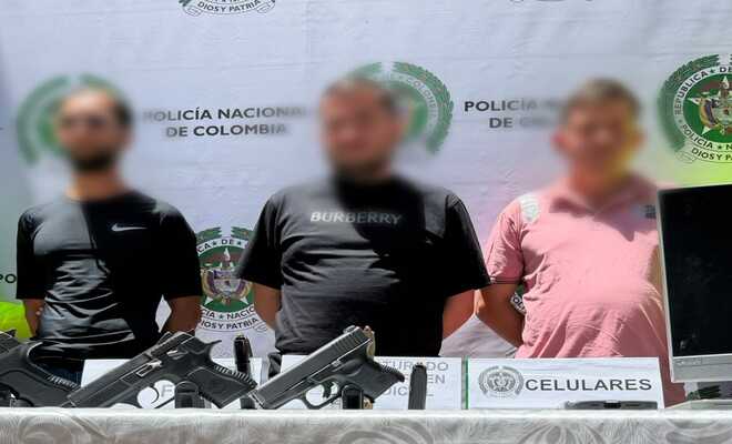 Tres personas de pie frente a un fondo institucional de la Policía Nacional de Colombia; en primer plano hay una mesa con dos armas cortas, un proveedor y un aviso identificado como “celulares”.