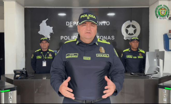 Un policía de pie realiza una entrevista frente a cámara detrás de él se observan dos policías de pie a los lados hay un computador y al fondo el escudo de la Policía Nacional y el mapa de Colombia