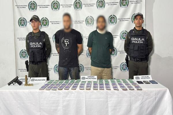 Un pend&oacute;n con escudo de la Polic&iacute;a Nacional, en pie cuatro personas dos uniformadas, una mesa con elementos, dinero, un arma y munici&oacute;n