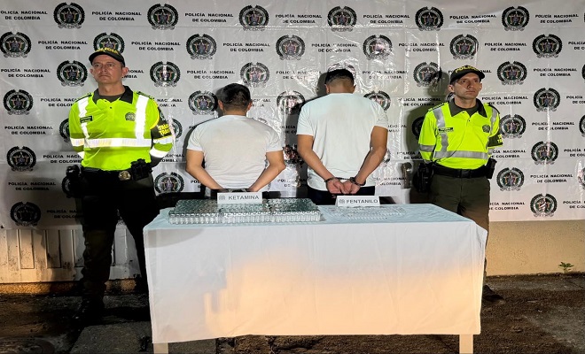 Dos personas de espaldas capturadas por el delito de tr&aacute;fico de sustancias para el procesamiento de narc&oacute;ticos, elementos incautados sobre una mesa con mantel blanco y dos Polic&iacute;as de la Seccional de Tr&aacute;nsito y Transporte brindando seguridad.
