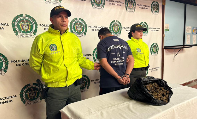 Hombre capturado en medio de dos Polic&iacute;as, en frente se encuentra ubicada una mesa con gramos de marihuana incautada