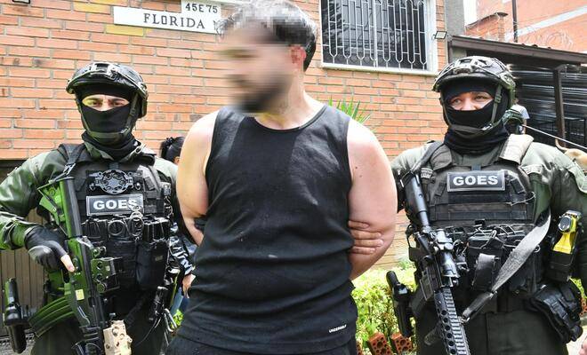 Exterior de edificio de ladrillo, en el centro hay una persona con camiseta negra y manos sujetas detr&aacute;s, acompa&ntilde;ada por dos personas uniformadas con equipo t&aacute;ctico que llevan chalecos con la palabra &ldquo;GOES&rdquo; y sostienen armas largas