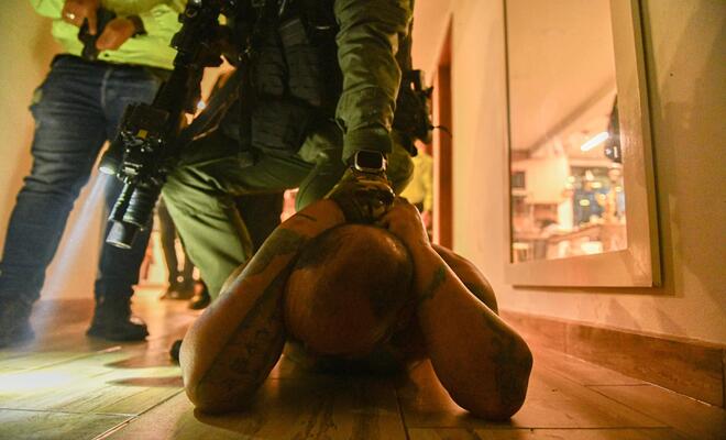  Interior con iluminaci&oacute;n c&aacute;lida donde una persona se encuentra acostada boca abajo sobre un piso de madera con las manos sujetas detr&aacute;s de la cabeza, mientras una persona uniformada se sit&uacute;a encima sosteniendo un arma larga; en el fondo se observan otras personas y un espacio tipo pasillo.