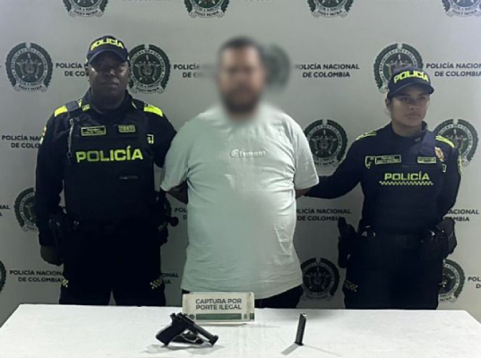fondo institucional de la Polic&iacute;a Nacional de Colombia; tres personas de pie, una en el centro con las manos sujetas detr&aacute;s, acompa&ntilde;ada por dos personas uniformadas a los lados; en primer plano hay una mesa con un arma corta y un objeto cil&iacute;ndrico, junto a un r&oacute;tulo que dice &ldquo;captura por porte ilegal"