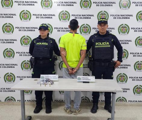 hombre capturado de camisa amarilla en medio de dos Policías, en frente una mesa blanca con arma de fuego incautada