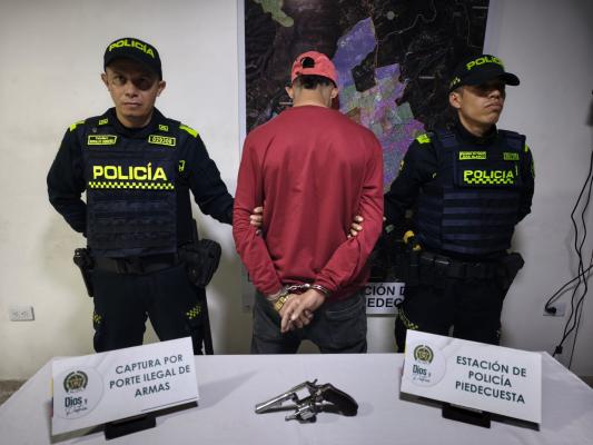 Hombre capturado con buso y gorra roja en medio de dos Policías, en frente sobre una mesa se encuentra el arma de fuego incautada