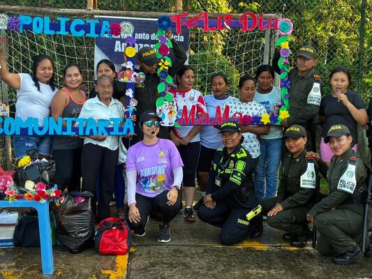 Un grupo mixto conformado por miembros de la fuerza policial y vecinos de la comunidad posan juntos de forma alegre y participativa, en un espacio al aire libre decorado con letreros y elementos coloridos que identifican la actividad. Se aprecian art&iacute;culos y materiales en el suelo, lo que evidencia que se trata de una jornada de integraci&oacute;n, convivencia y acercamiento entre la instituci&oacute;n y la poblaci&oacute;n
