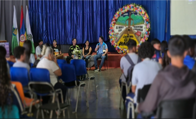 Auditorio con personas sentadas frente a un escenario donde se desarrolla un conversatorio; varias personas participan en un panel junto a una persona uniformada, con una decoración floral de gran tamaño y banderas institucionales al fondo