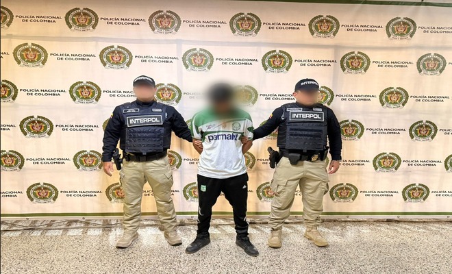 En un espacio institucional, una persona con el rostro difuminado se encuentra de pie entre dos integrantes de la Policía Nacional que portan chalecos con la identificación de INTERPOL. Al fondo se observa un panel con los logos repetidos de la Policía Nacional de Colombia.