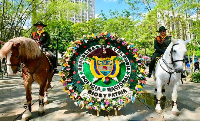 Estructura floral circular con el escudo Policía Nacional ubicada entre dos caballos montados por personal uniformado en un espacio público arborizado.